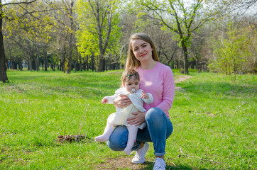 Fototapeta premium Mom with baby in bright clothes on a pink plaid on the green right. Family resting in the park on a warm day. Mom and little girl 10 months walk in the park