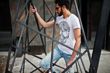 Street style arab man in eyeglasses with longboard posed inside metal pyramid construction.