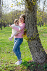 Fototapeta premium Mom with baby in bright clothes on a pink plaid on the green right. Family resting in the park on a warm day. Mom and little girl 10 months walk in the park