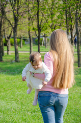 Fototapeta premium Mom with baby in bright clothes on a pink plaid on the green right. Family resting in the park on a warm day. Mom and little girl 10 months walk in the park