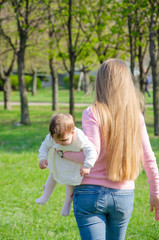 Fototapeta premium Mom with baby in bright clothes on a pink plaid on the green right. Family resting in the park on a warm day. Mom and little girl 10 months walk in the park