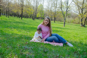 Mom with baby in bright clothes on a pink plaid on the green right. Family resting in the park on a warm day. Mom and little girl 10 months walk in the park