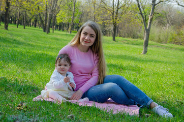 Fototapeta premium Mom with baby in bright clothes on a pink plaid on the green right. Family resting in the park on a warm day. Mom and little girl 10 months walk in the park