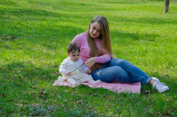 Fototapeta premium Mom with baby in bright clothes on a pink plaid on the green right. Family resting in the park on a warm day. Mom and little girl 10 months walk in the park