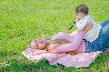 Mom with baby in bright clothes on a pink plaid on the green right. Family resting in the park on a warm day. Mom and little girl 10 months walk in the park