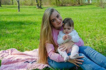 Mom with baby in bright clothes on a pink plaid on the green right. Family resting in the park on a warm day. Mom and little girl 10 months walk in the park