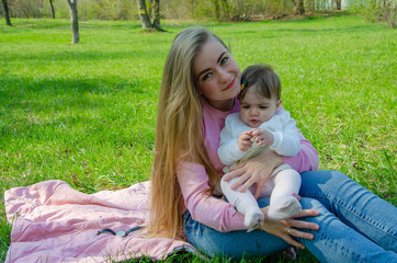 Mom with baby in bright clothes on a pink plaid on the green right. Family resting in the park on a warm day. Mom and little girl 10 months walk in the park