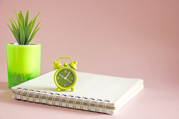 The alarm clock in green shows the time of 10 o'clock in the afternoon, a notebook with white sheets is next, an office flower,copy space. Working time, time to work, Office background
