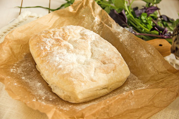 Ciabatta italian white bread on parchment paper on a wooden table, next to fresh greens and cotton napkin. View from above