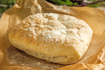 Ciabatta italian white bread on parchment paper on a wooden table, next to fresh greens and cotton napkin.