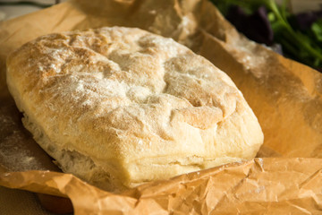 Ciabatta italian white bread on parchment paper on a wooden table, next to fresh greens and cotton napkin.