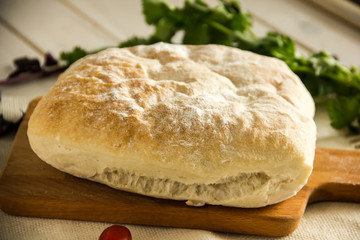Ciabatta italian white bread on parchment paper on a wooden table, next to fresh greens and cotton napkin.