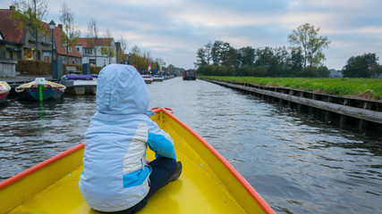 A boy with a blue jacket with his back on the front of the boat on the water canals in Giethoorn, the Netherlands, full of boats and water.