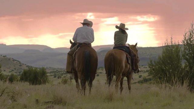 Cowboy and cowgirl stop for a rest sitting on horses, view of the mountain range and sunset ahead, 29 fps.
