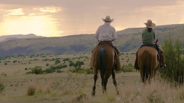 Vintage style, husband and wife ranchers watch the sunrise on horseback, mountain range ahead, slow motion 24 fps.