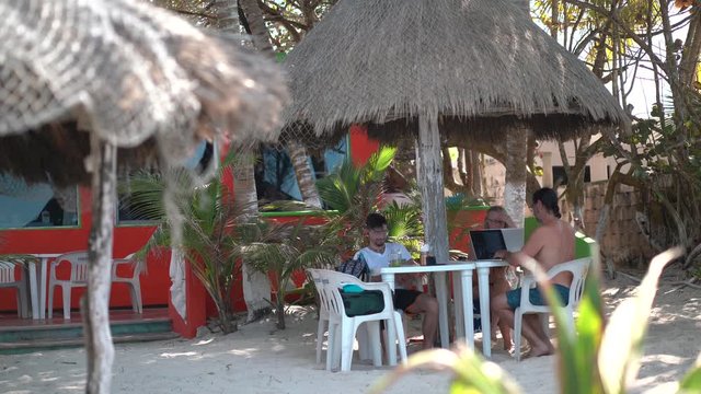 Mature mother and father with teenage son sitting under a palapa at a table outside at a beach resort working on computers and smart phone.