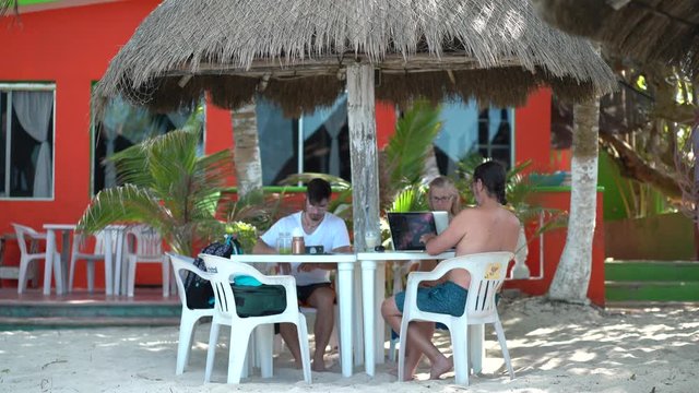 Mature mother and father with teenage son sitting under a palapa at a table outside at a beach resort working on computers and smart phone.