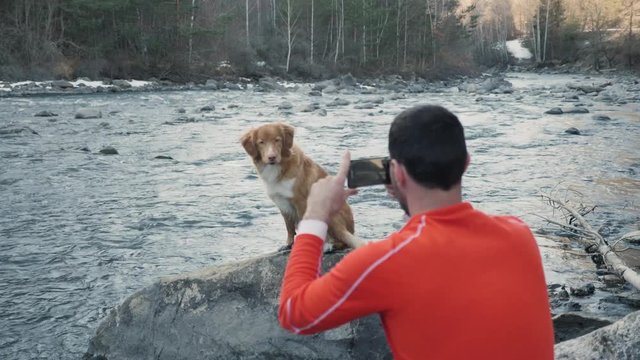 A man taking a picture of his pet golden Scottish Retriever dog next to a river while on a nature hike in slow motion and showing love and affection.