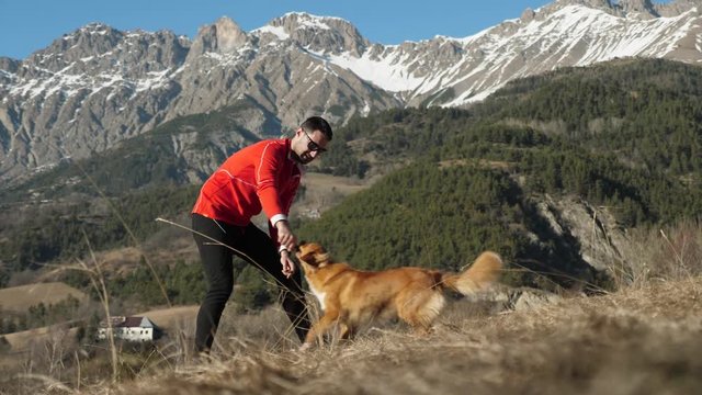 A man in a red shirt and his pet Scottish Retriever dog play fetch together with a stick in slow motion with the snowy French alps mountains in the background.