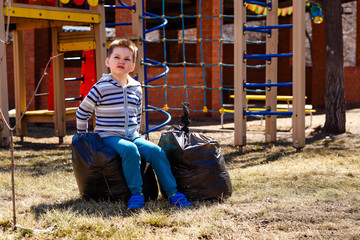 five-year-old boy sitting on a large plastic bags with garbage on the Playground . Spring cleaning of the environment from garbage