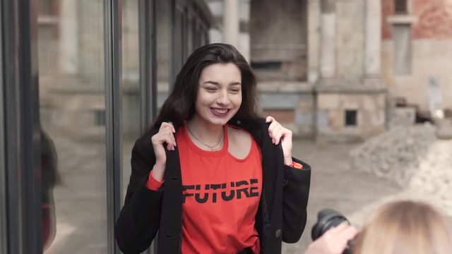 Young female photographer is making a photo of a young beautiful female model who is posing outside near the building with a glass wall.