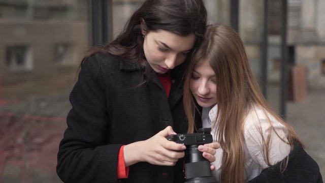 Two cute girls are looking at camera's screen on the photos they have made and discuss it. They are standing outside.