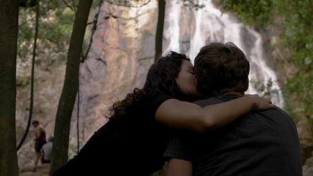 A young couple sit cuddling and kissing, admiring the view of a majestic, rocky waterfall