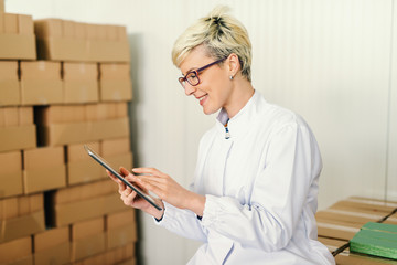 Happy Caucasian blonde employee suing tablet for work while sitting around boxes in food factory.