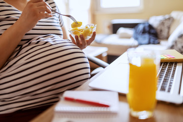Young Caucasian pregnant freelancer eating fruits and sitting at home office. On desk laptop, glass with fresh orange juice and notebooks. Working pregnant women concept.