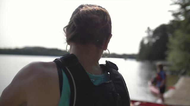 A young woman standing on the edge of a lake puts on sunscreen as she gets ready to go out on the water.