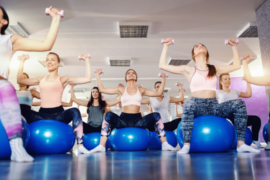 Group Of Focused And Inspired Group Of Sporty Women Sitting On Pilates Ball And Lifting Dumbbells In Gym. Small Changes Can Make A Big Difference.