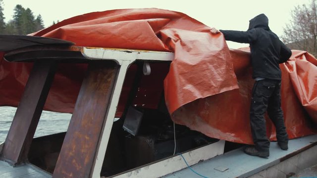 Young man untying  waterproof cover of wooden boat before starting restoration maintenance