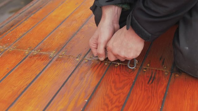 Young man uses pocket knife to clean out old mastic of wooden boat roof planking close up