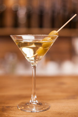 Delicious martini drink with green olives on stick over a wooden bar counter - obrazy, fototapety, plakaty