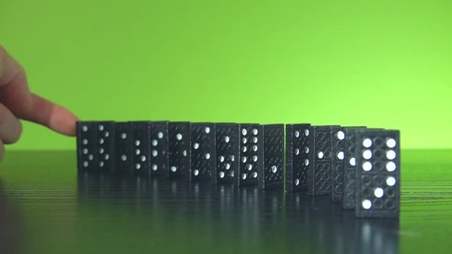 Slow motion shot of a finger nudge some black dominoes which stands in a half circle line for a chain reaction in front of a green background with plenty copy space for text.