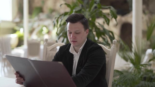 Young good-looking man in a white shirt and black jacket is sitting in a huge expensive restaurant, looking through the menu and talking.