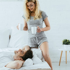 cheerful blonde girl holding cups with coffee near happy man lying in bed