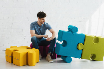 cheerful man looking at connected blue and green jigsaw pieces while sitting near brick wall
