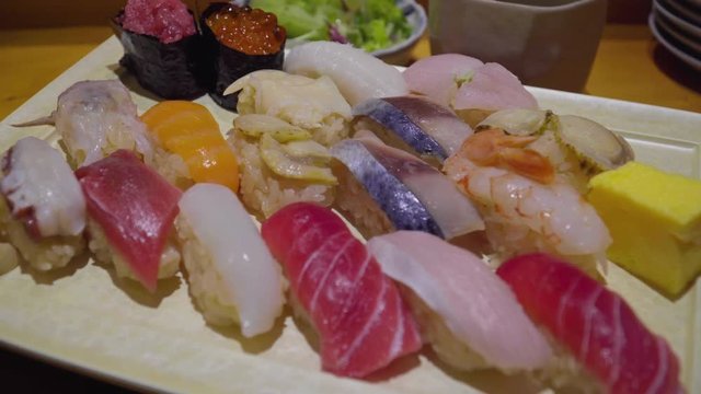 medium pan down food shot of a set plate of different japanese sushi specialties, nigiri, maki and sashimi on a white rectangle pate and wooden table, indoor setting