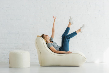 cheerful blonde girl lying on  bean bag chair and gesturing near brick wall