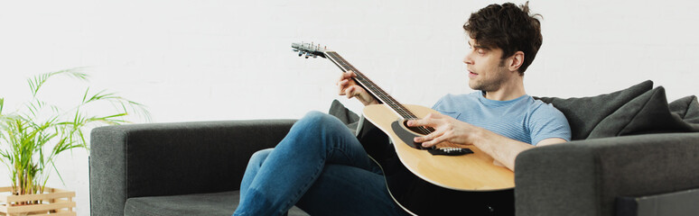 panoramic shot of handsome man sitting on sofa and playing acoustic guitar at home