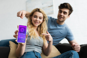 selective focus of cheerful man and blonde woman pointing with fingers at smartphone with shopping app on screen