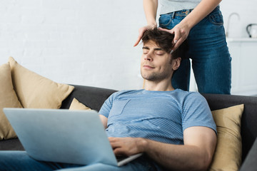 cropped view of woman touching head of happy man sitting on sofa with laptop