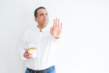 Middle age man eating asian noodles from take away carton with open hand doing stop sign with serious and confident expression, defense gesture