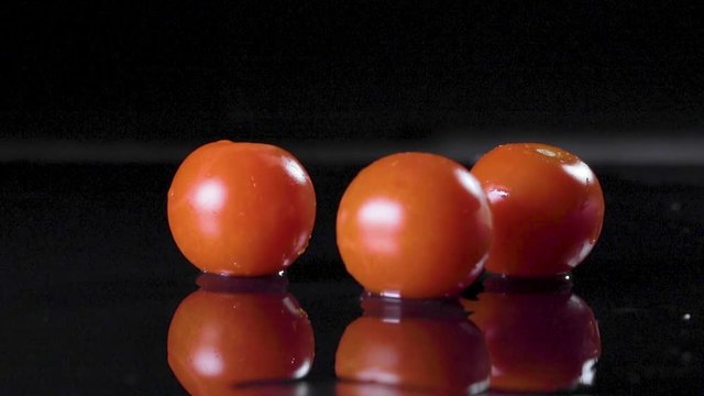 Pomodorini rossi che cadono su un piano bagnato con sfondo nero e schizzano acqua ovunque. Ripresa al rallentatore.