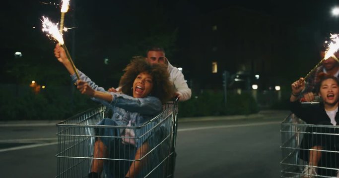 Slow motion of group of young friends of different ethnicities are having fun together racing on shopping carts and sparklers at supermarket parking at night.