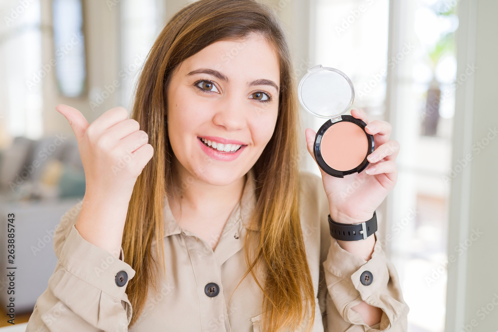 Sticker Beautiful young woman using make up cosmetics applying powder pointing and showing with thumb up to the side with happy face smiling