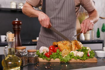 Hands of chief man cuting baked chicken with vegetables with a big knife on a wooden plate in a restaurant