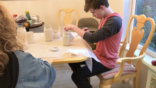 Teenage boy smiling while painting mason jars with white paint for turning them into vases at an upcoming event