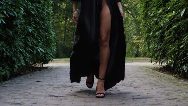 Medium shot of beautiful girl walking and wearing an open black dress, showing her tanned legs and high heels. Surrounded by trees, walking on a pathway.